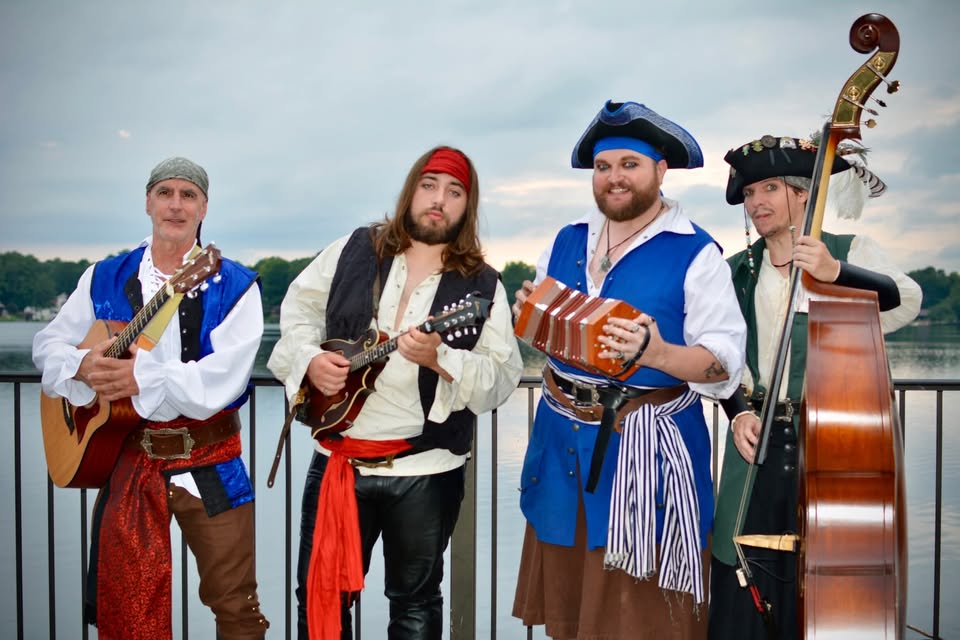 530828126_764267853024842_5070396502584705010_n Four musicians in pirate attire with instruments by a waterfront.