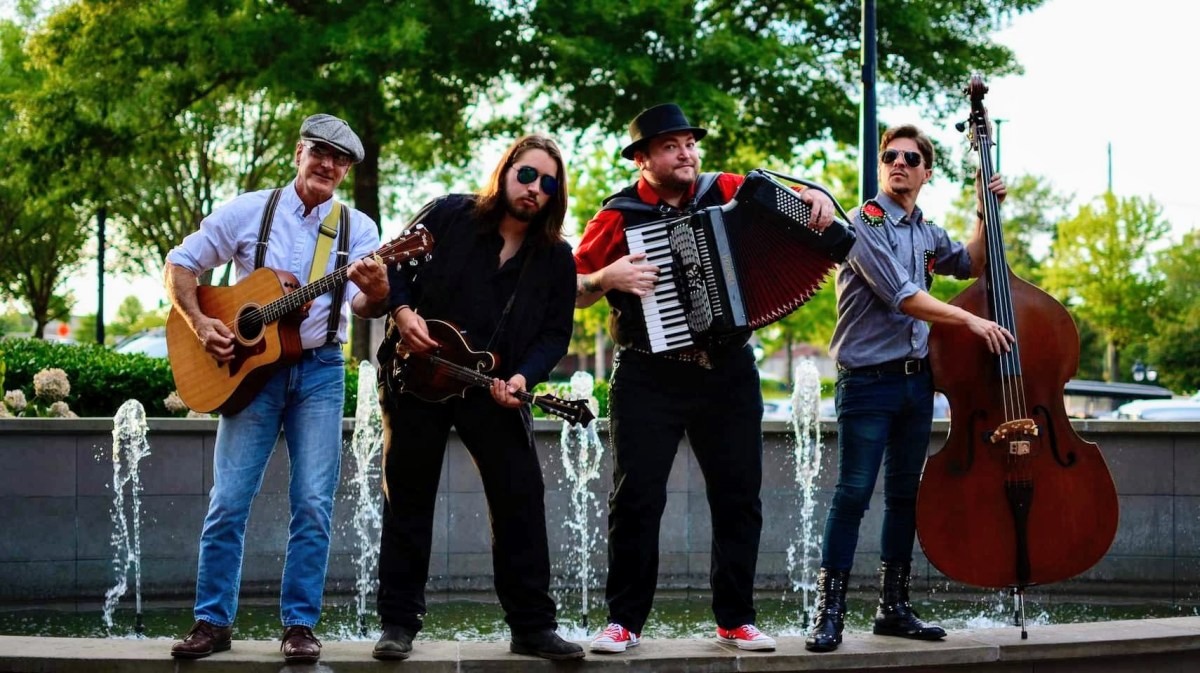 Kraken’s Release Band Pic (2) Four musicians with instruments standing in front of a fountain outdoors.